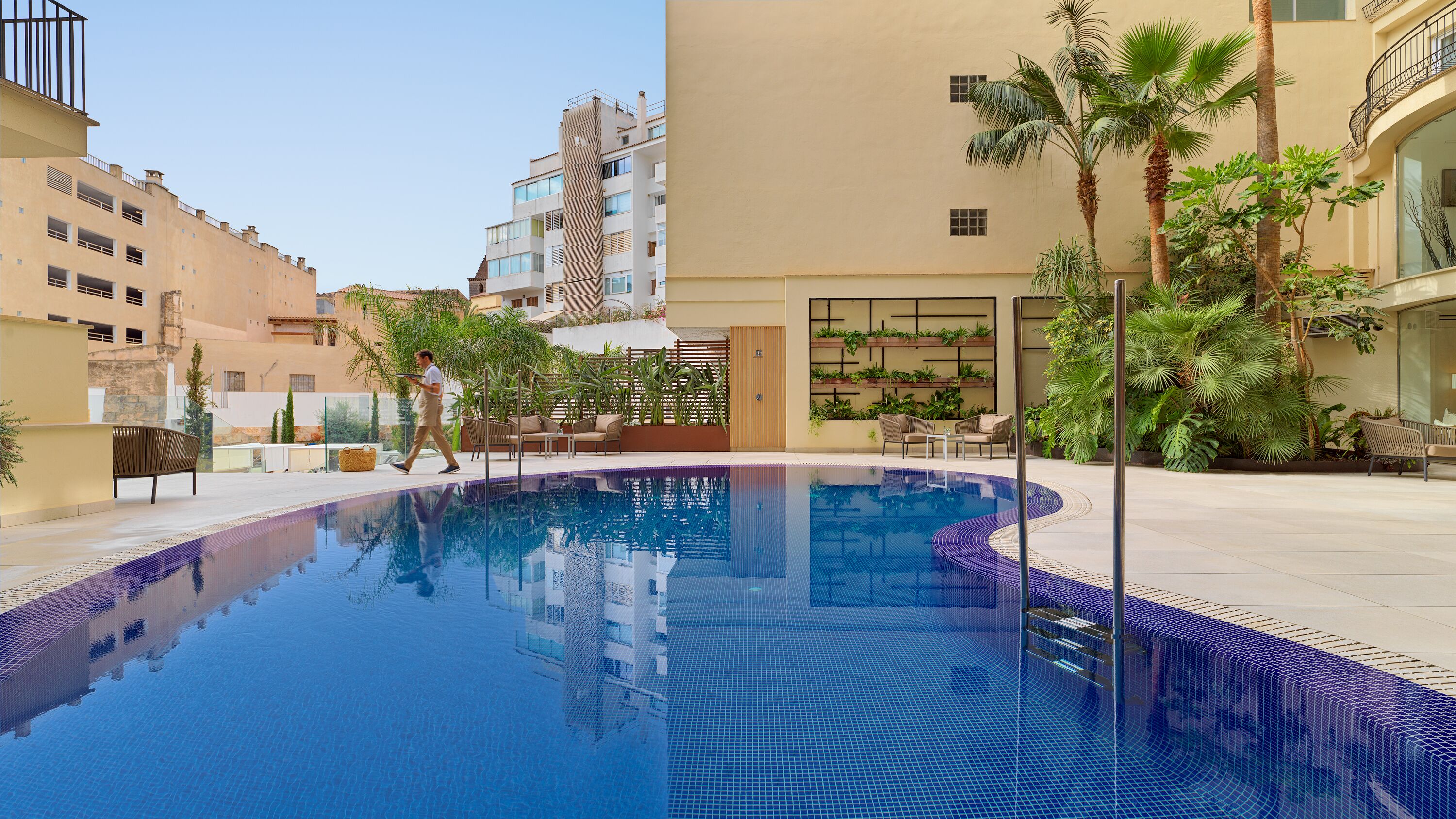 Una piscina con borde de azulejos, rodeada de mobiliario exterior y palmeras.