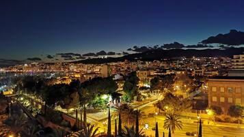Un paisaje urbano nocturno con edificios iluminados, un puerto deportivo y un telón de fondo montañoso.