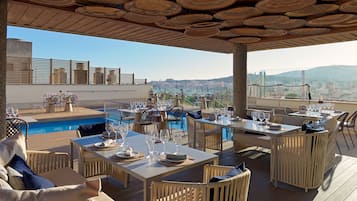 Un área de comedor al aire libre con mesas y sillas, una piscina y vistas a un paisaje urbano.