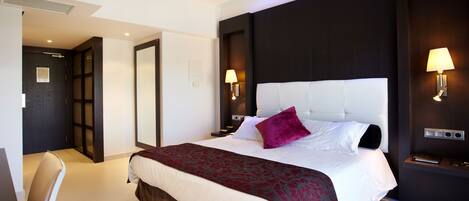 Una habitación de hotel moderna con una cama grande, un escritorio y una silla. Se observa una puerta que conduce a otra habitación y una lámpara fijada a la pared.