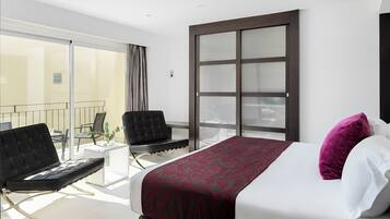 A modern hotel room with a large bed, a black leather chair, a small table, and a balcony with a glass door.