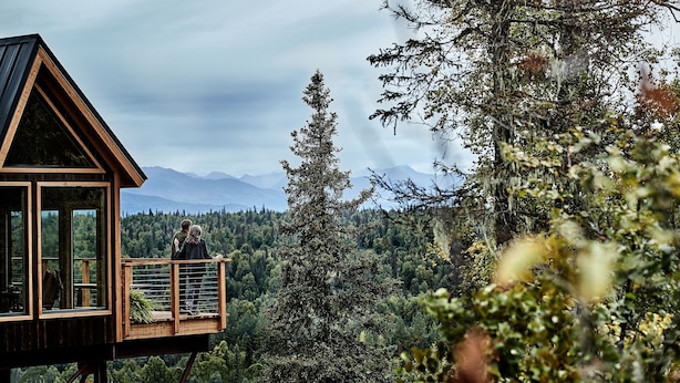 Una cabaña de madera con balcón que da a un paisaje boscoso y montañas distantes.