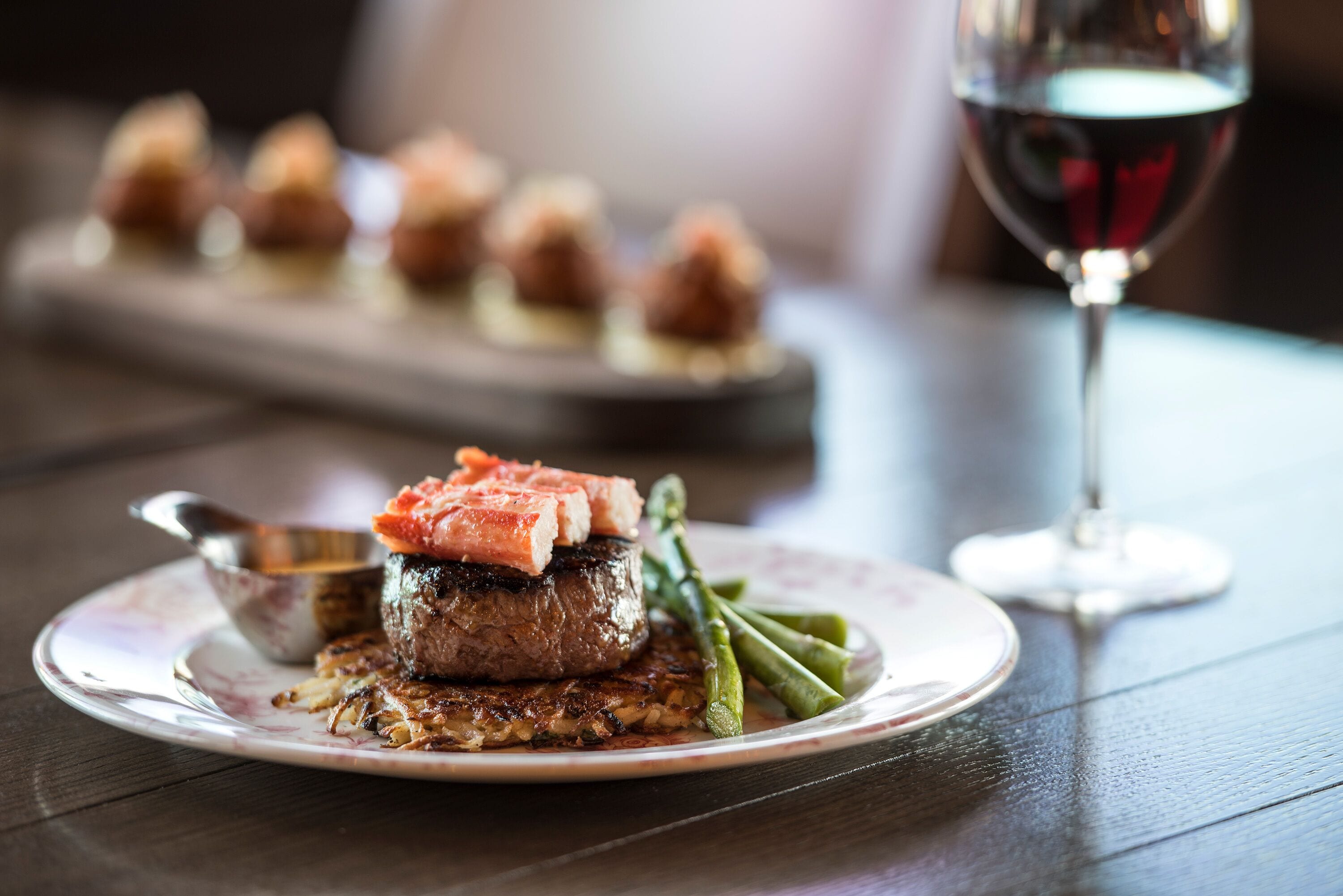A plate with a steak topped with salmon, asparagus, and a small bowl of sauce.