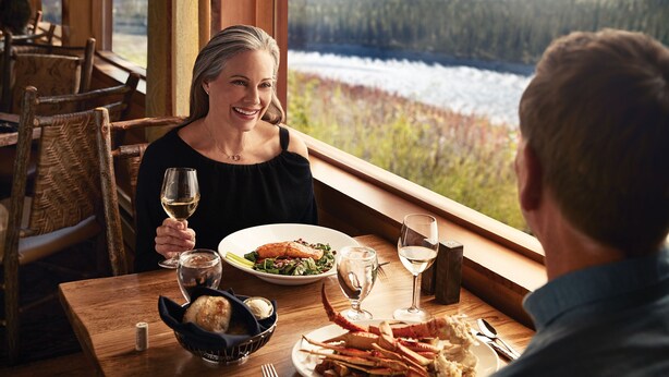 A dining table with a meal, wine glasses, and a scenic view of a forest and river. - Denali Princess Wilderness Lodge (Denali National Park)