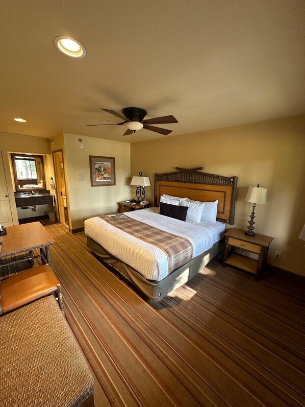 A hotel room with a bed, a desk, a chair, a ceiling fan, and a nightstand with a lamp.