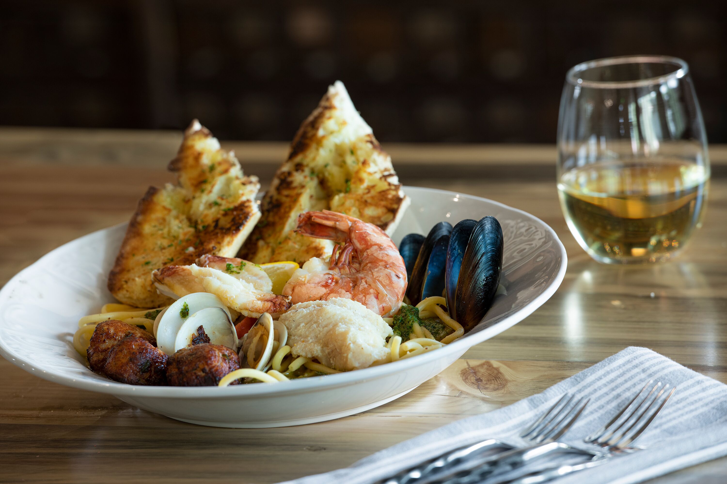 A plate of seafood pasta with bread and a glass of white wine on a wooden table.
