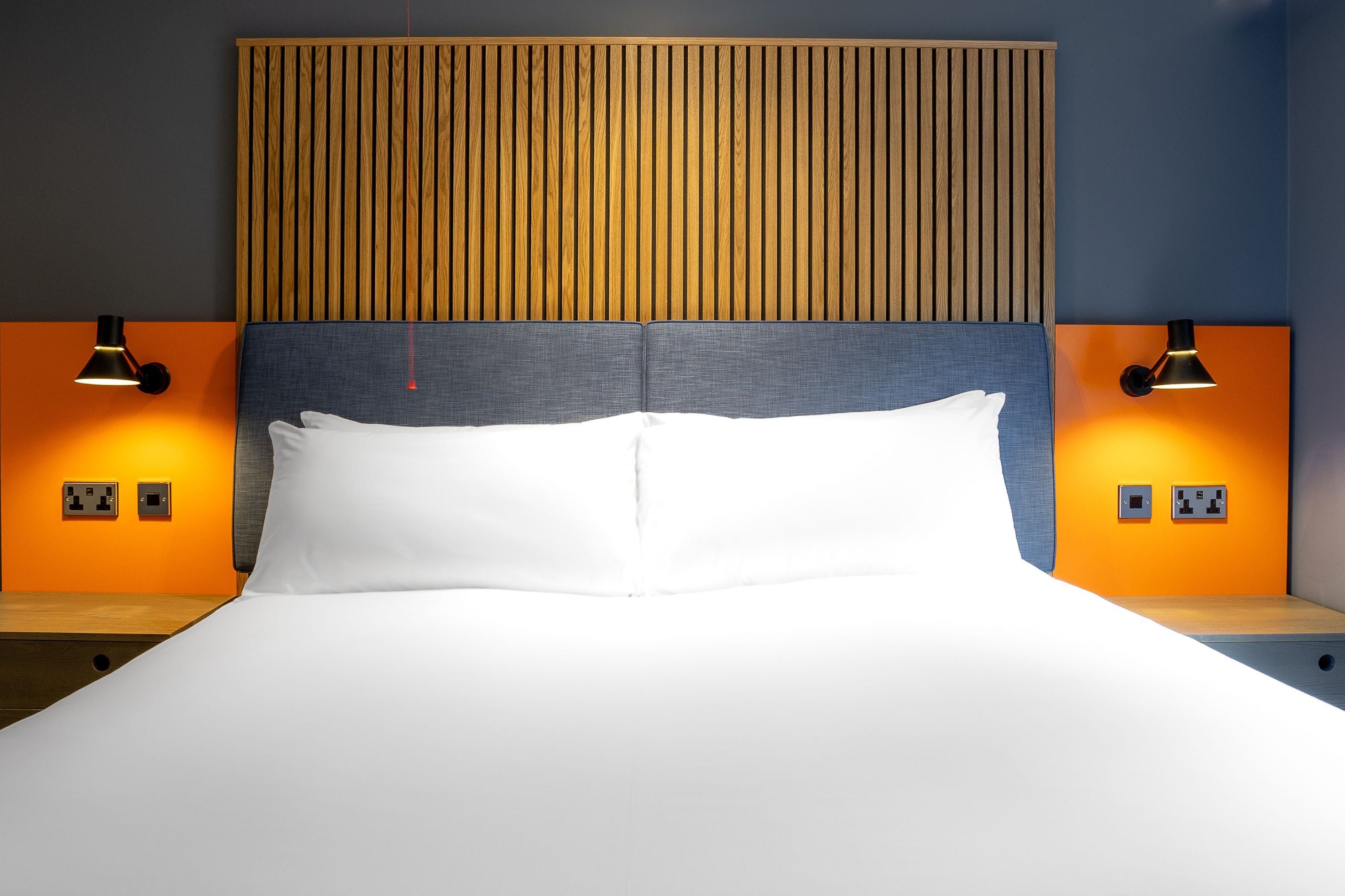 A hotel room with a bed, two bedside lamps, a wooden headboard, and a wall socket.