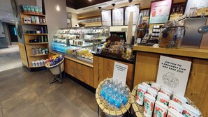 A coffee shop interior with a display case of pastries, a coffee machine, and a sign promoting reusable cups. - Village Hotel Leeds North (Leeds)