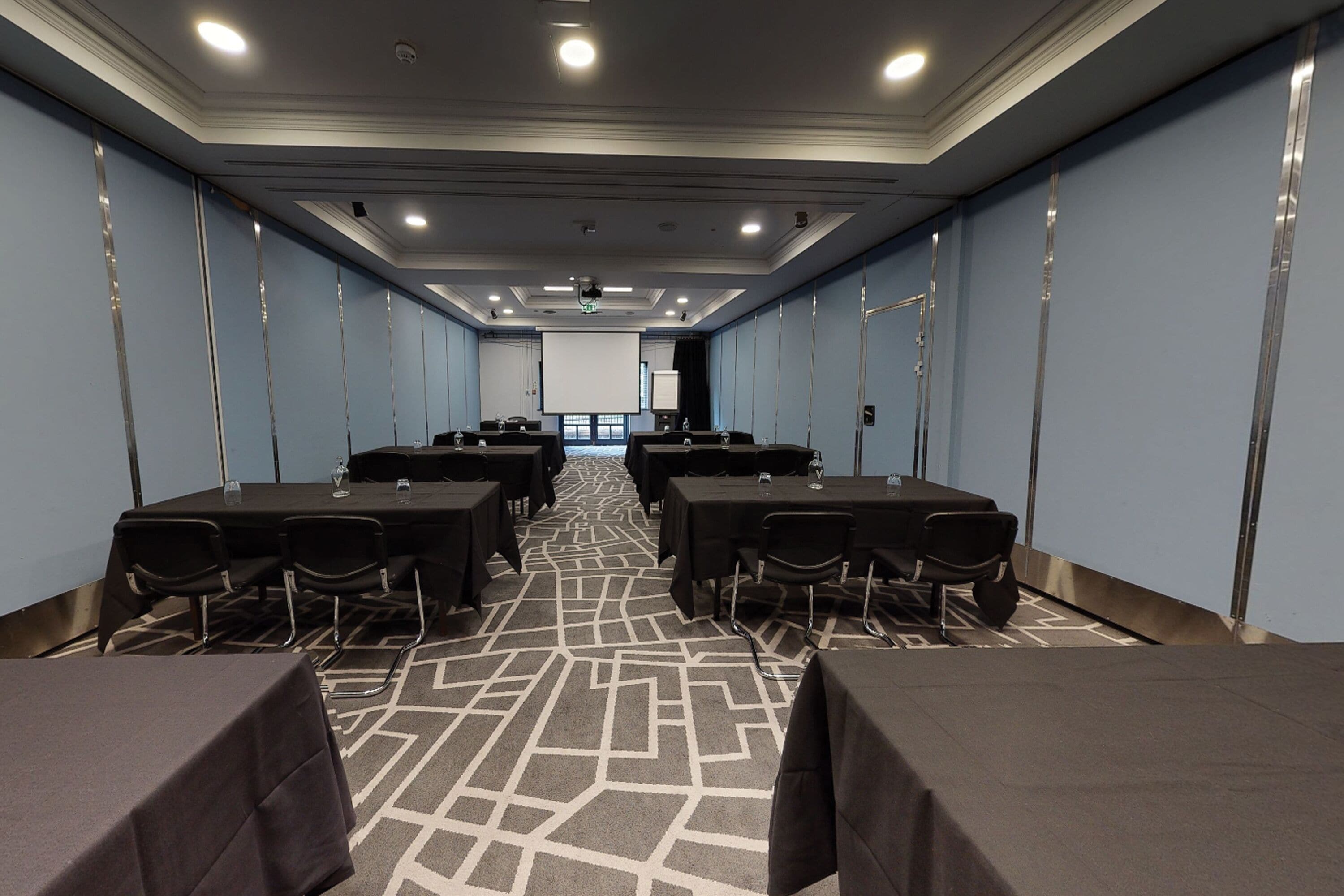 Una sala de conferencias con mesas y sillas, una pantalla de proyector y una alfombra estampada.