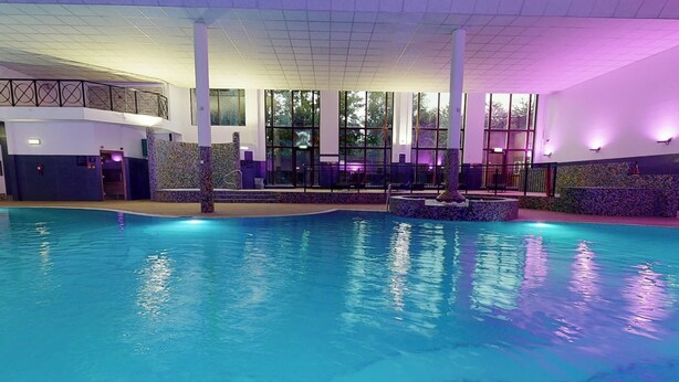 An indoor swimming pool with clear water, surrounded by walls with large windows and a balcony with a railing. - Village Hotel Leeds North (Leeds)