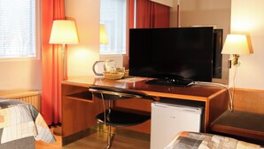 A hotel room with a wooden desk, a television, a mini-fridge, a chair, a bed with a patterned blanket, and a window with curtains. - Hotel Sommelo (Kouvola)
