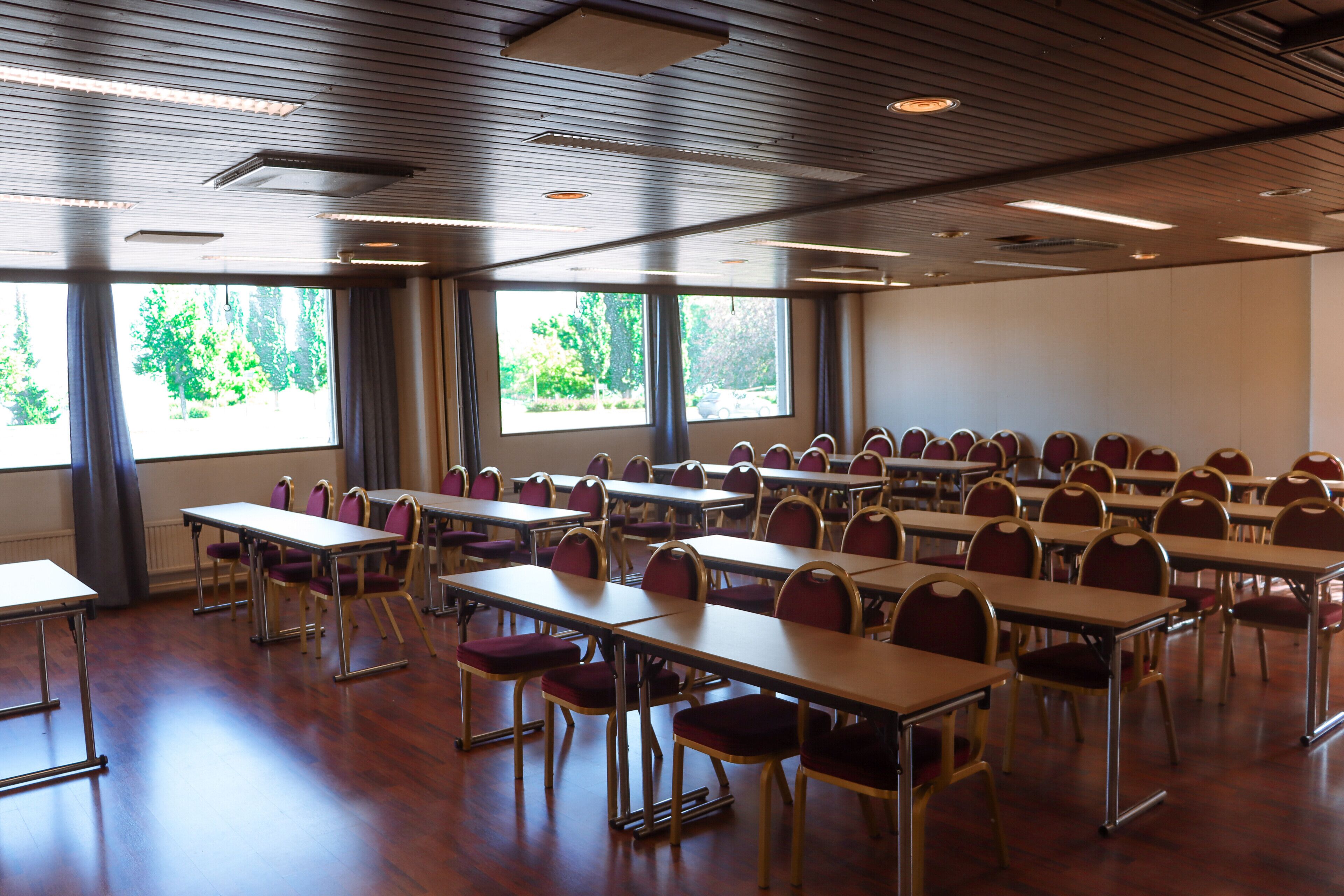A conference room with rows of tables and chairs, large windows with a view of trees, and a wooden ceiling.