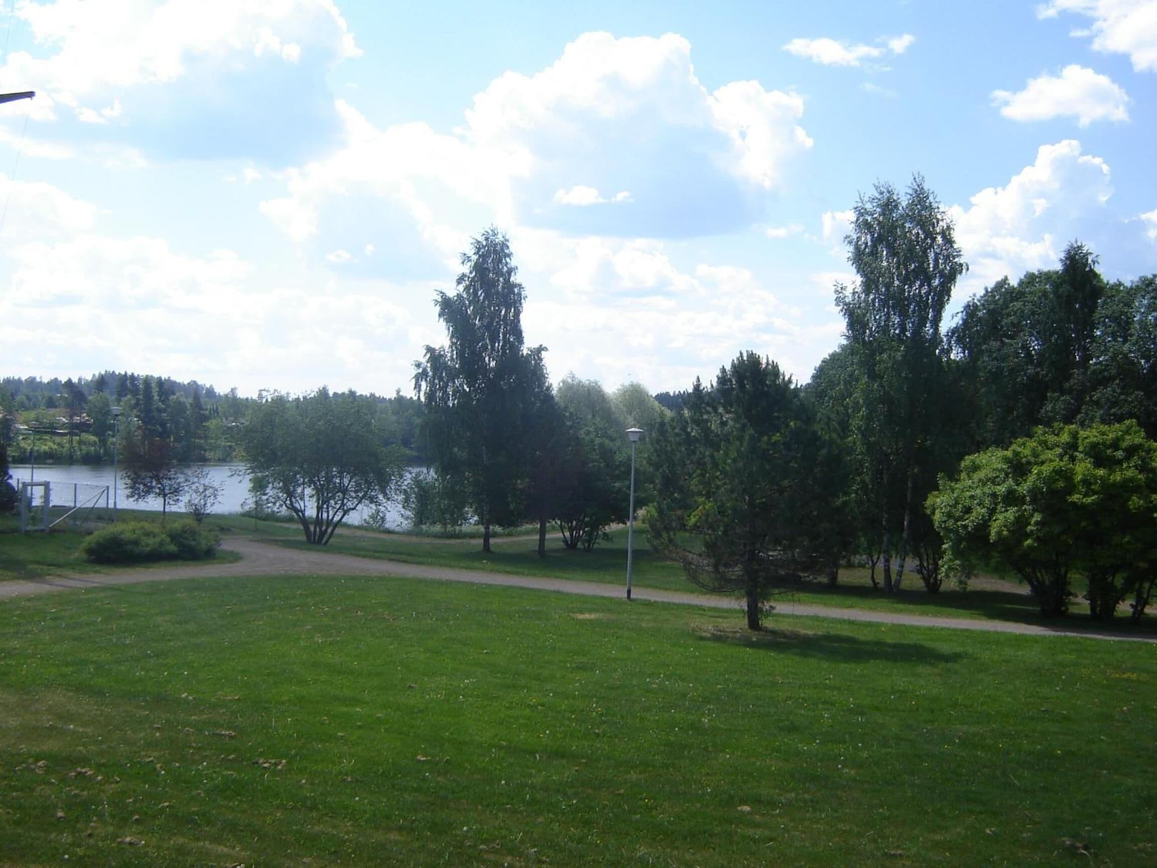 A park with a grassy field, trees, a path, and a body of water in the background.