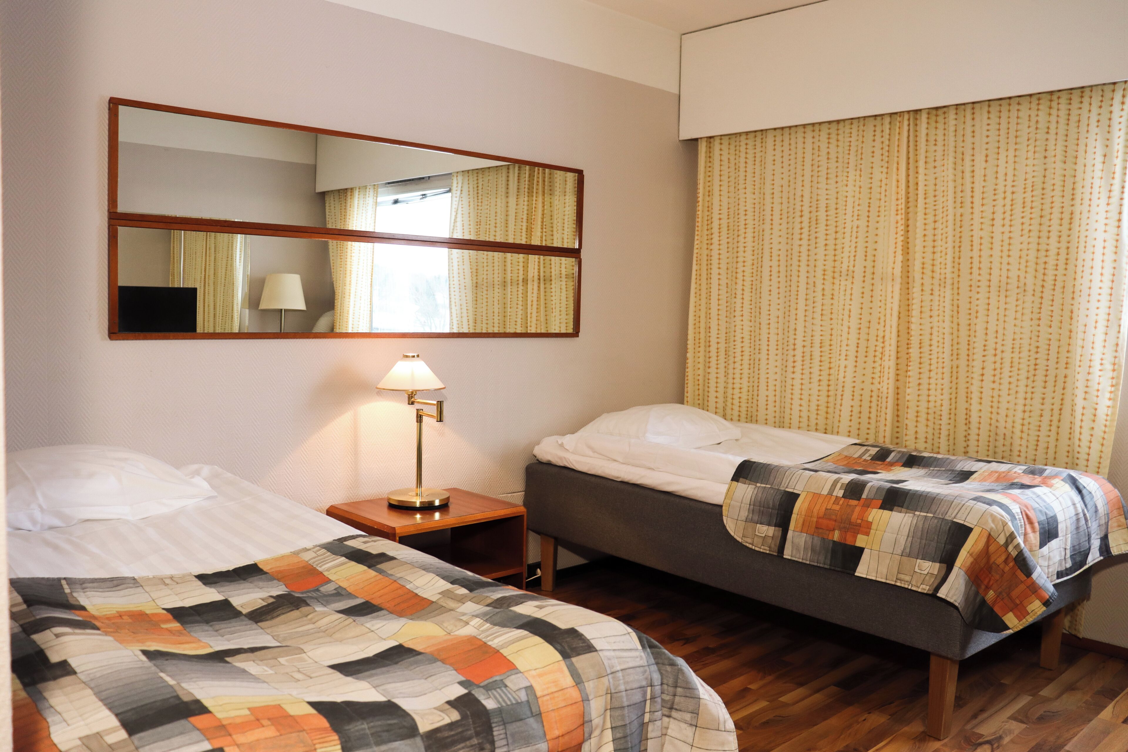 A hotel room with two beds, a nightstand with a lamp, a large mirror, and a window with curtains.