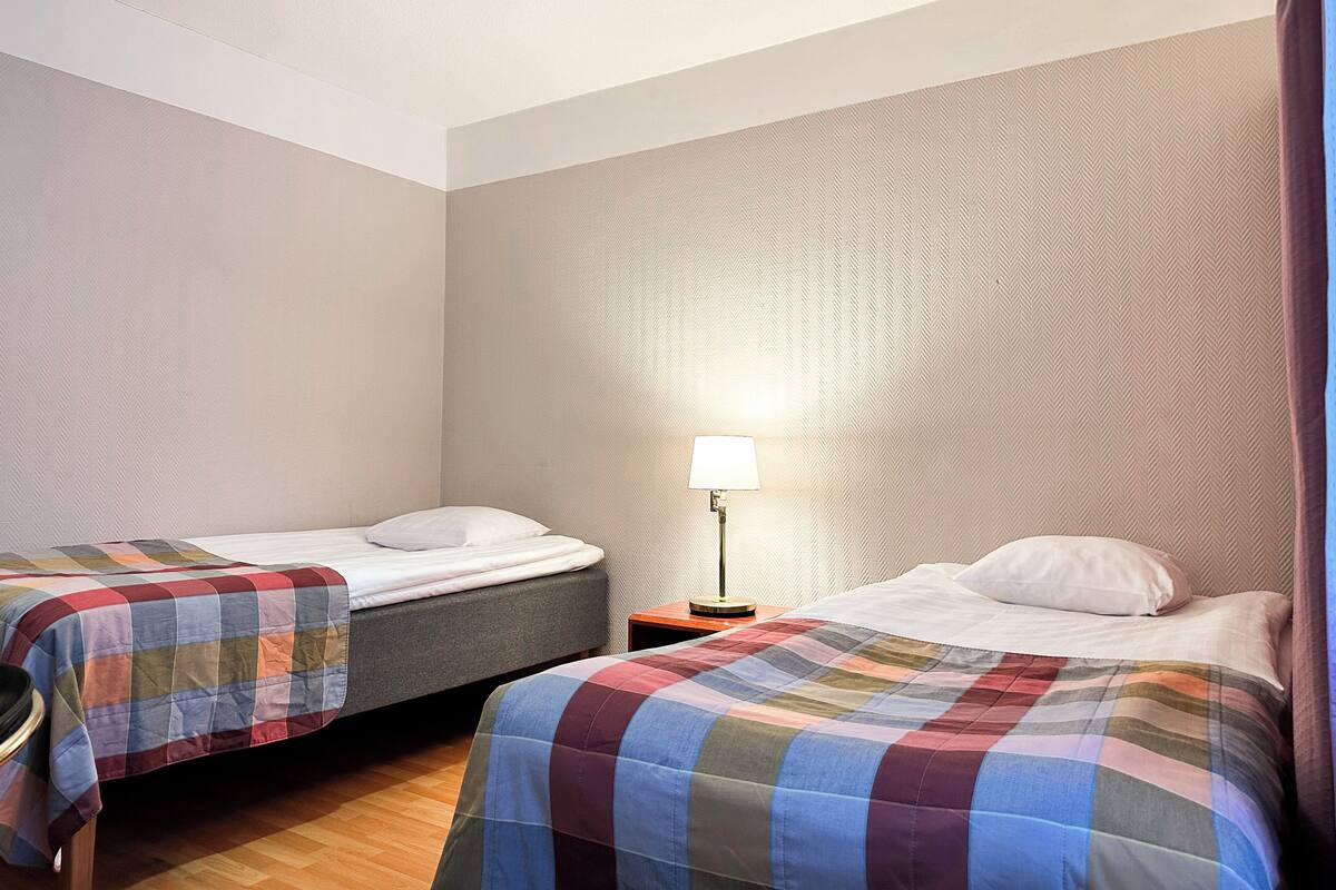 A hotel room with two single beds, a nightstand with a lamp, and a patterned wall.