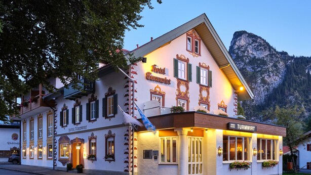 Un hotel europeo tradicional con un letrero que dice “Hotel Turnwirt” y “Zimmerhäuser”. Presenta un estilo arquitectónico clásico con el paisaje de montañas al fondo.