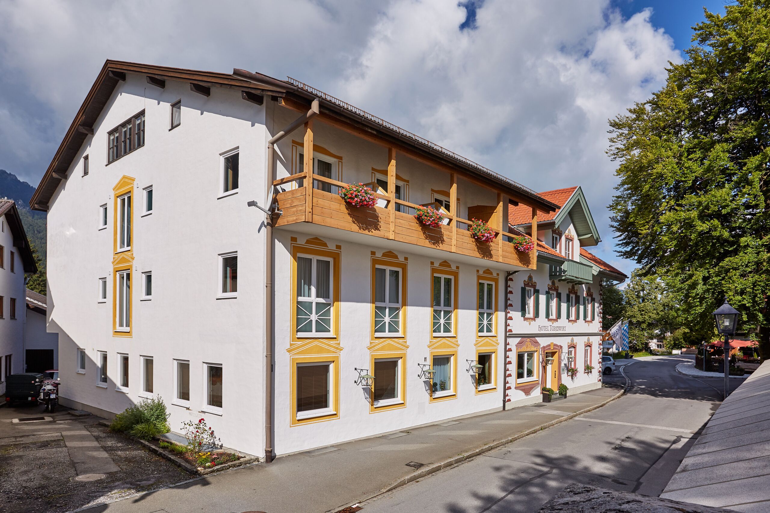Ein zweigeschossiges Gebäude mit Balkon und Blumenkörben, an einer Straße mit parkendem Auto und Baum.