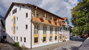 Ein zweigeschossiges Gebäude mit Balkon und Blumenkörben, an einer Straße mit parkendem Auto und Baum.