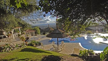 Zona de piscina con sombrilla de paja, mobiliario exterior y vistas al paisaje.