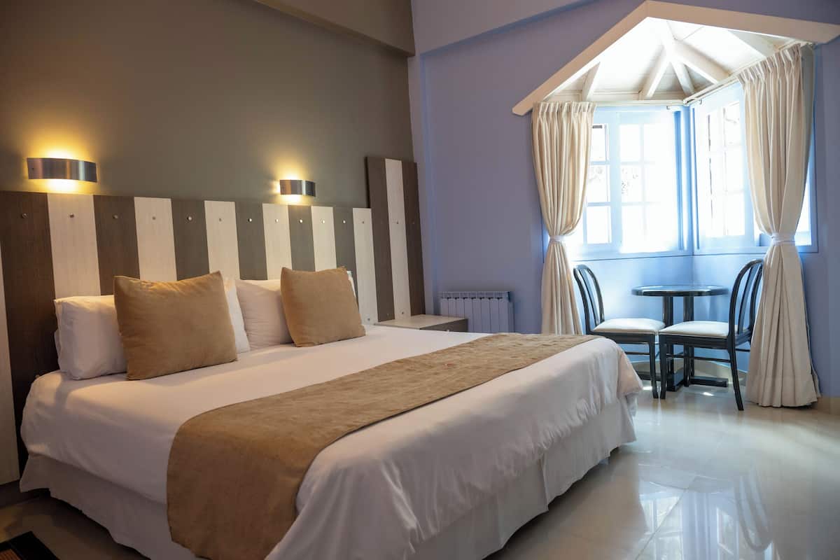 A hotel room with a large bed, two bedside lamps, a window with curtains, and a small table with chairs.