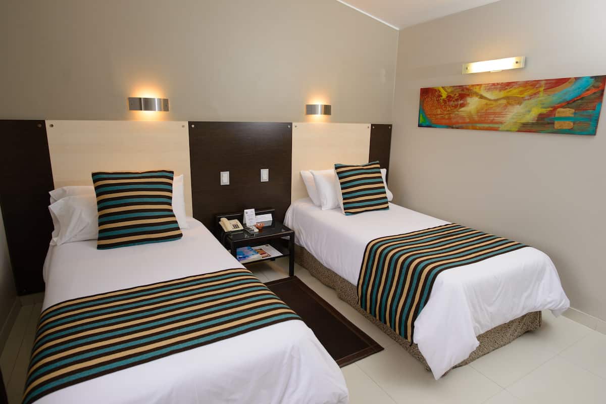 A hotel room with two beds, each with striped bedding and pillows, a nightstand with a phone and a magazine, and a painting on the wall.