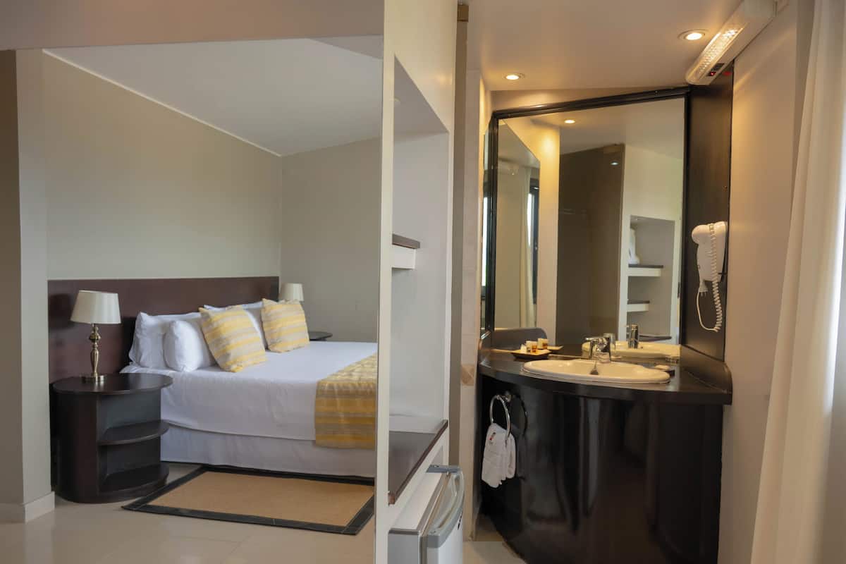 A hotel room with a bed, bedside table, lamp, and a large mirror above the sink.