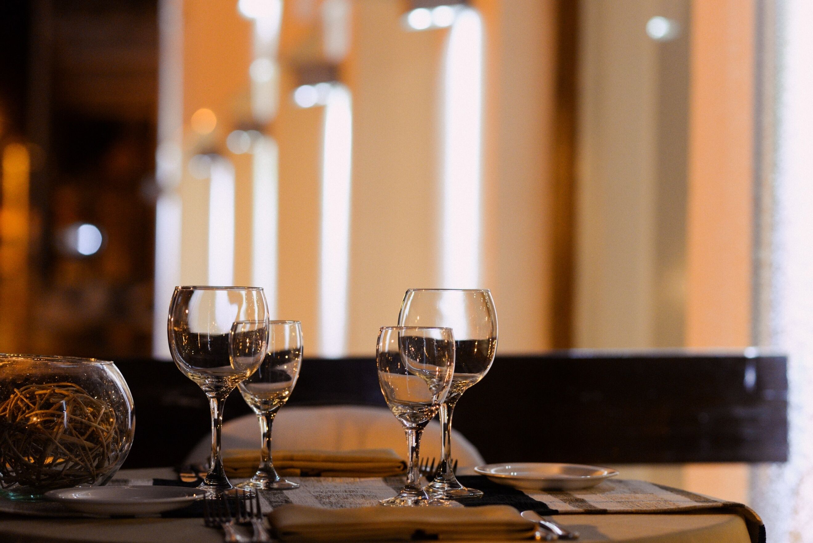 A table set for dining with three wine glasses and a glass bowl, in a dimly lit room with vertical light fixtures.