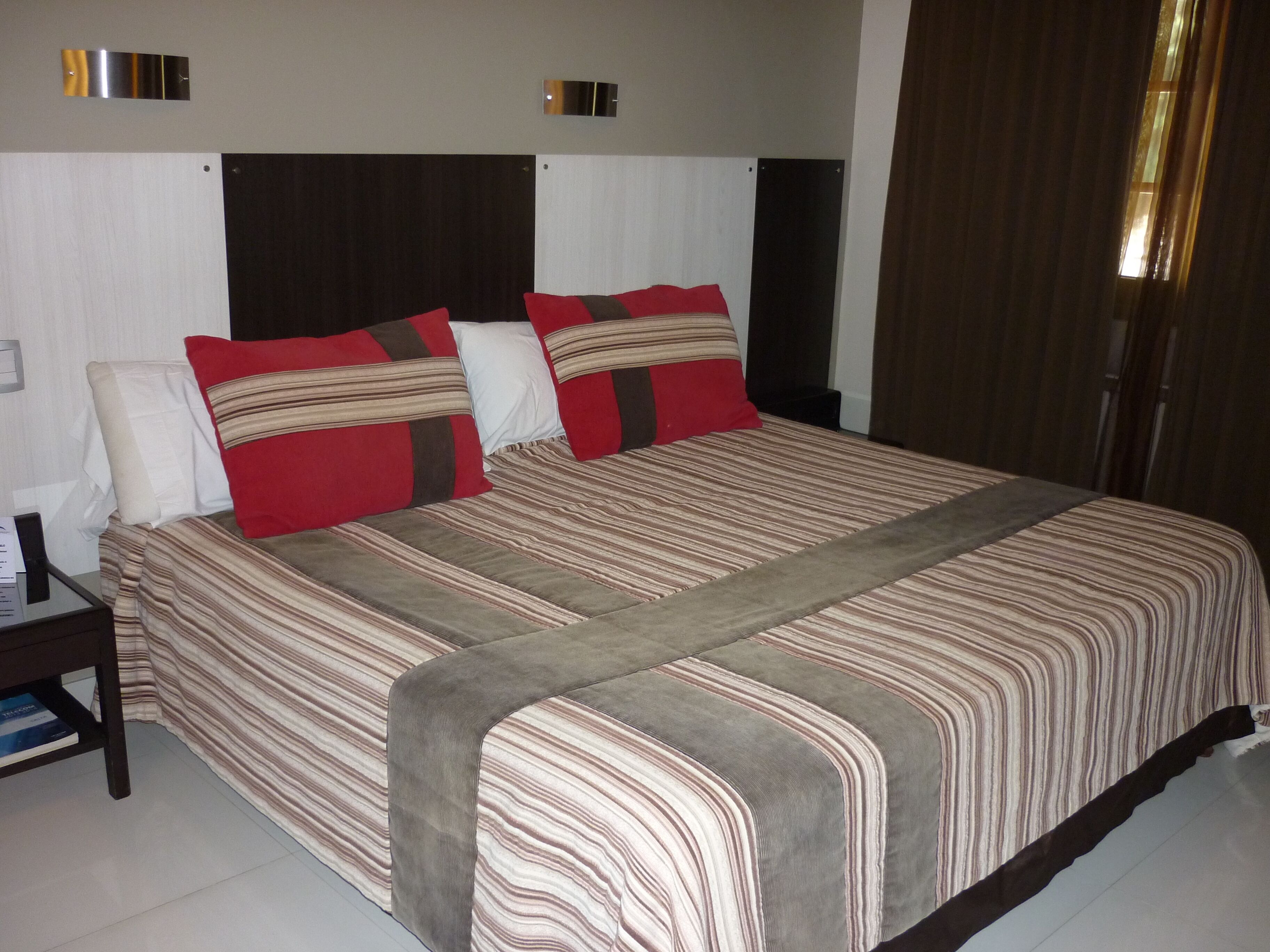 A neatly made bed with striped bedding and two pillows, a bedside table with a book, and a window with curtains.