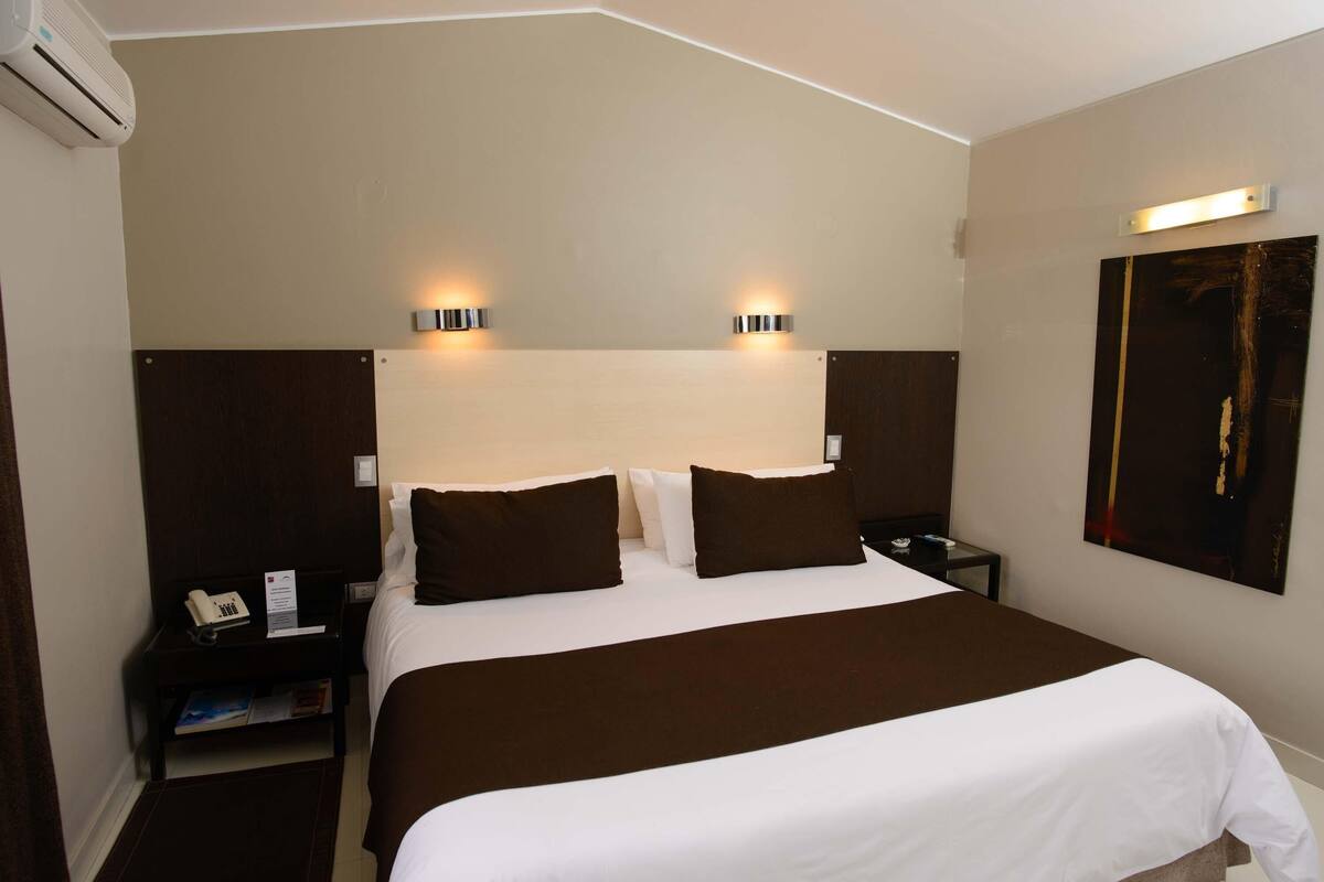 A hotel room with a large bed, two bedside tables, an air conditioning unit, and a wall-mounted artwork.