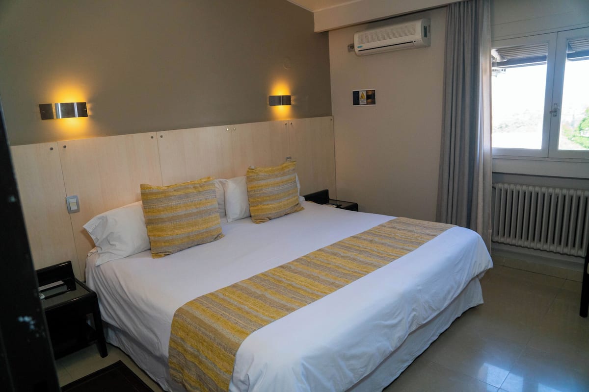 A hotel room with a large bed, two bedside tables, a nightstand, a window with curtains, and an air conditioning unit on the wall.