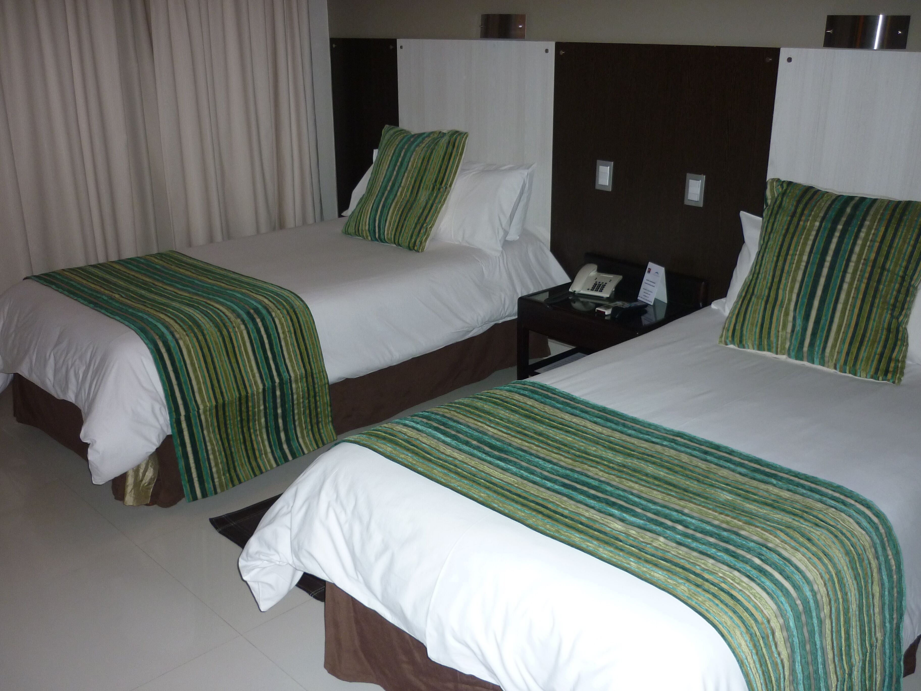 Two double beds with green-striped bedspreads, a bedside table with a phone, and a curtain in the background.