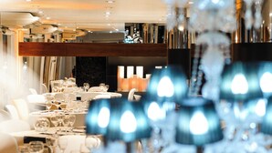 Une salle à manger bien éclairée, avec des tables dressées pour le repas, un lustre moderne et un plafond haut doté d’un éclairage encastré.