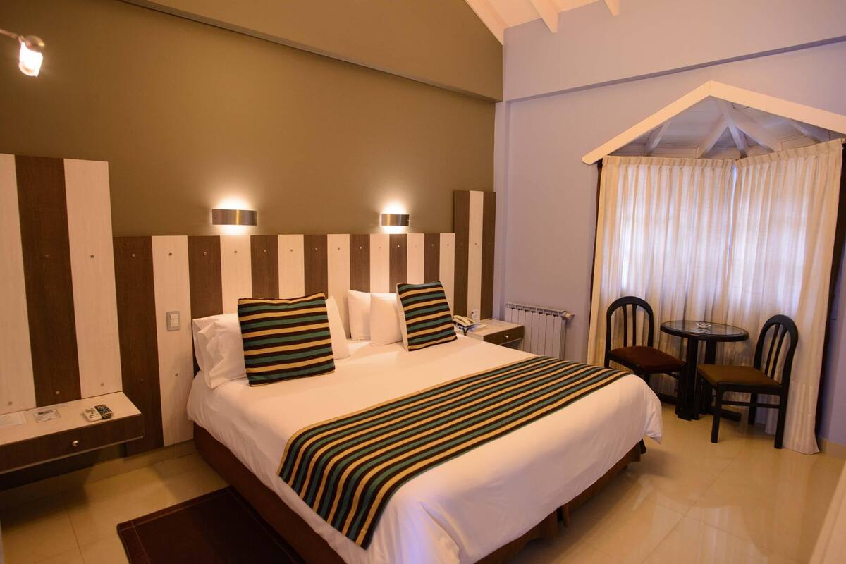 A hotel room with a large bed, bedside tables, a small table with chairs, and a window with curtains.