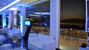Un espace lounge moderne avec des canapés blancs, un bar et une vue sur la ville la nuit.