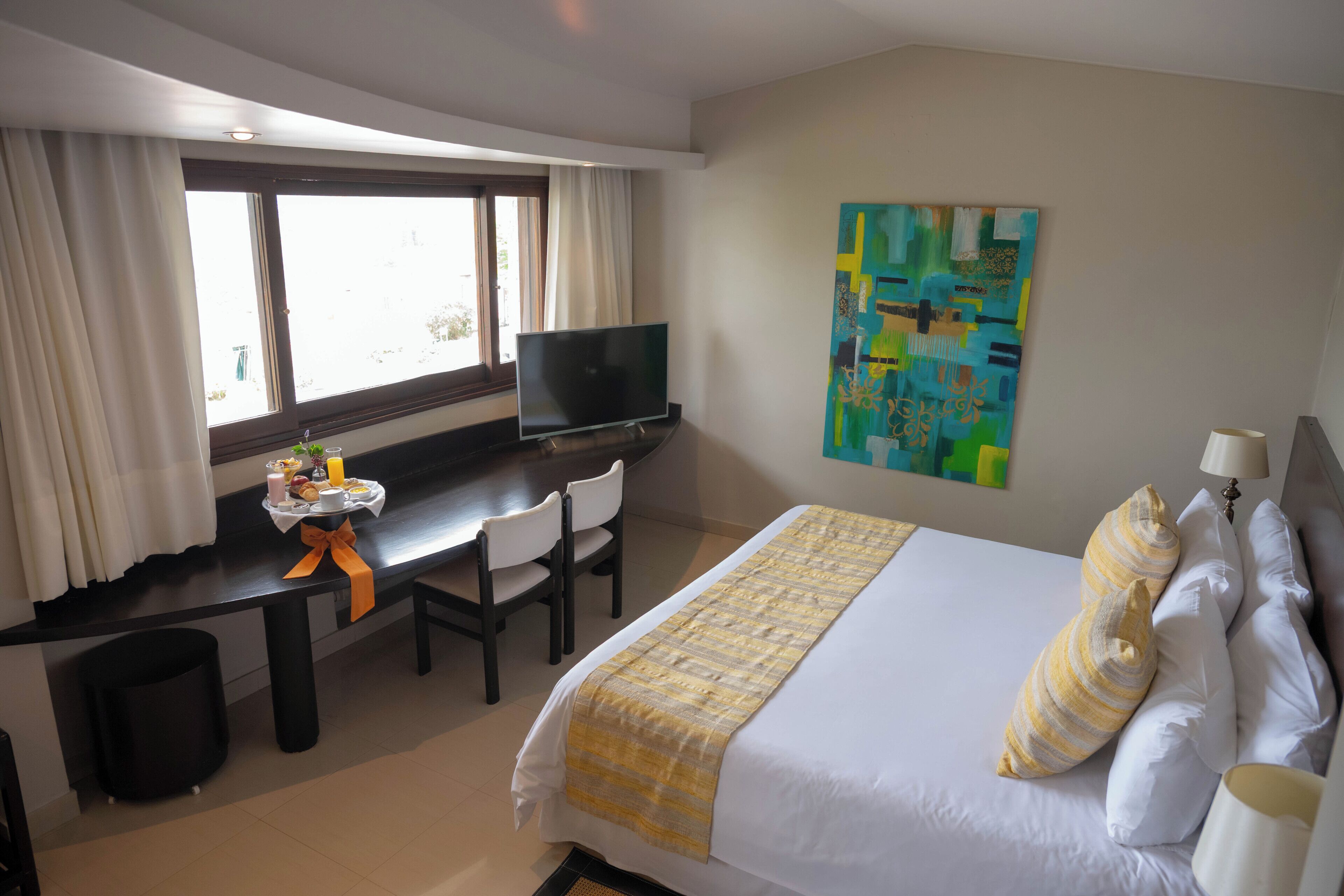 A hotel room with a bed, a desk with a TV, a window with curtains, and a painting on the wall.