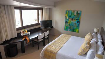 A hotel room with a bed, a desk with a TV, a window with curtains, and a painting on the wall.