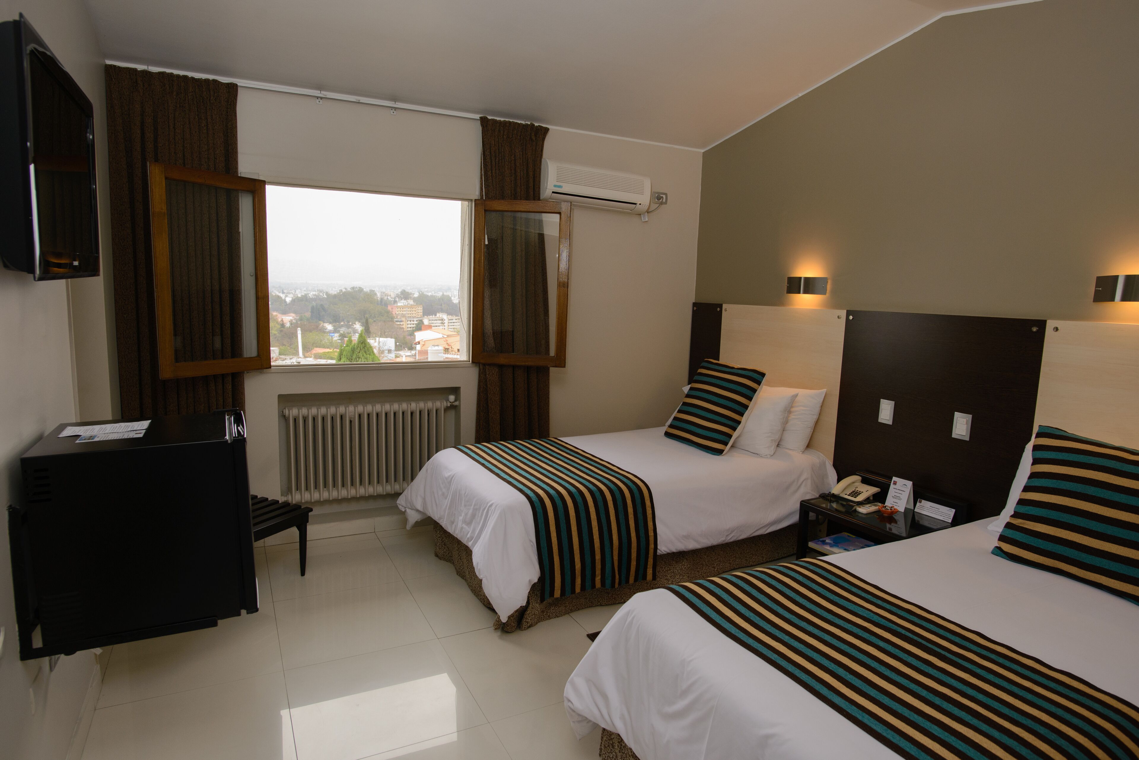 A hotel room with two beds, a desk, a chair, a radiator, and a window with curtains.