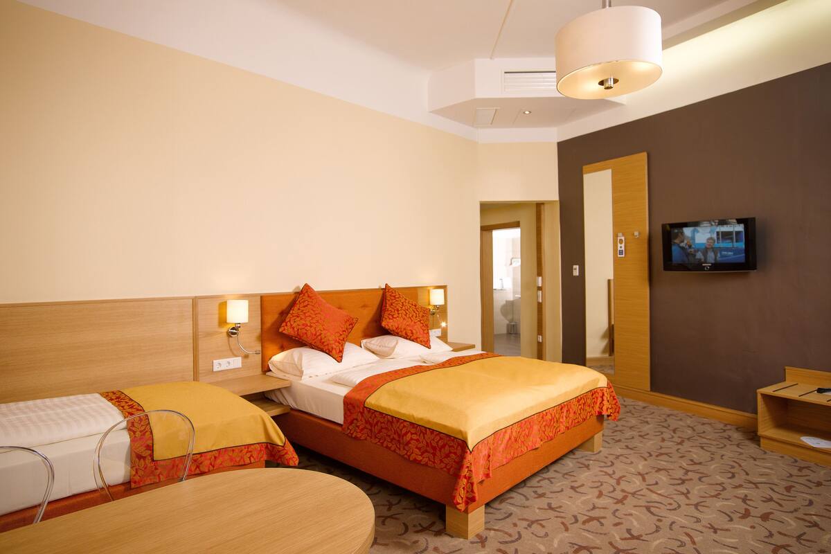 Habitación de hotel con una cama grande, una cama más pequeña, una mesa redonda y un televisor en la pared.