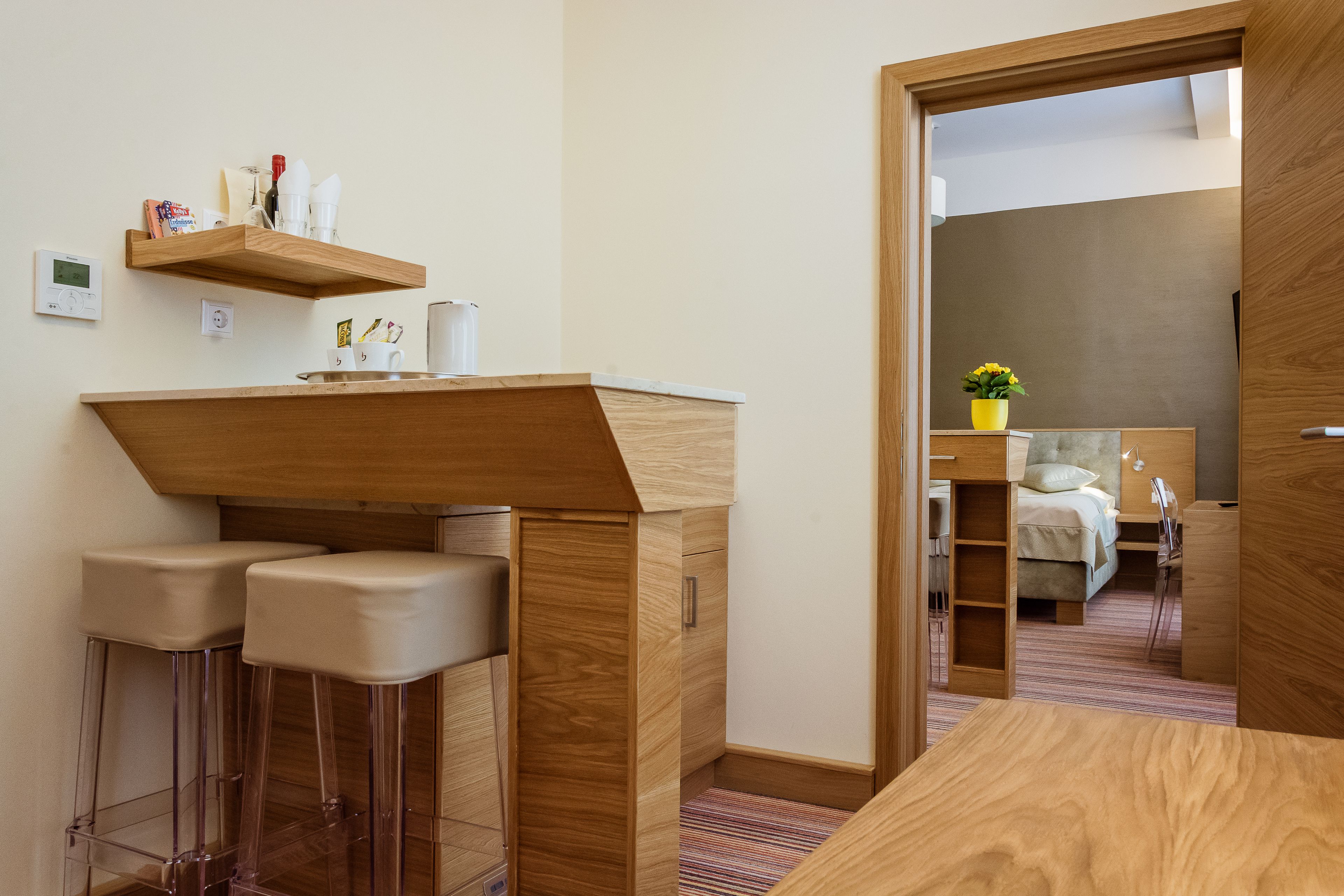 Une pièce de taille modeste, avec un bureau en bois, deux tabourets de bar, une petite étagère contenant des articles de toilette et un lit avec une table de chevet en arrière-plan.