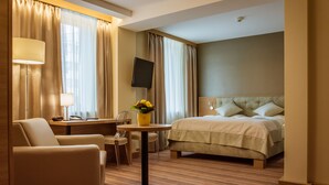 A hotel room with a bed, a desk, a chair, a table with a vase of flowers, and a TV on the wall.