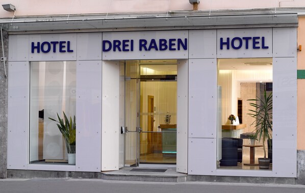 A hotel entrance with glass doors, a potted plant, and a visible interior with a sofa and a table.