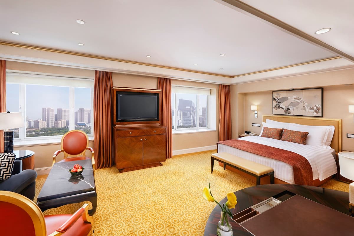A hotel room with a bed, a TV, a desk, a chair, a bench, and a view of the city.