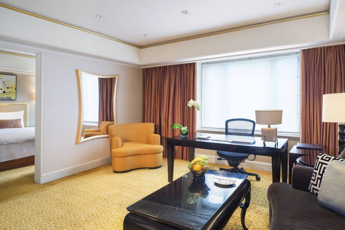 A hotel room with a bed, a sofa, a desk with a lamp, a chair, and a large window with curtains.