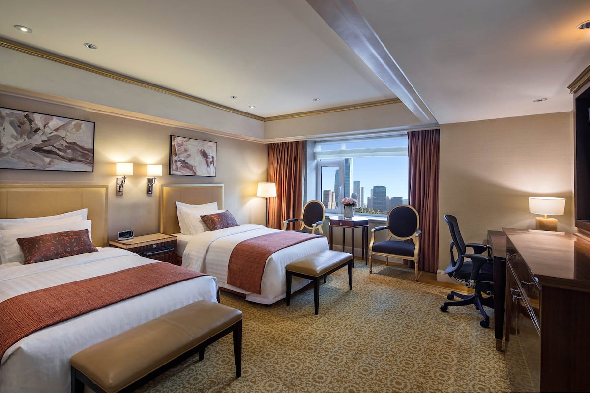 A hotel room with two beds, a desk, a chair, and a view of the city skyline.