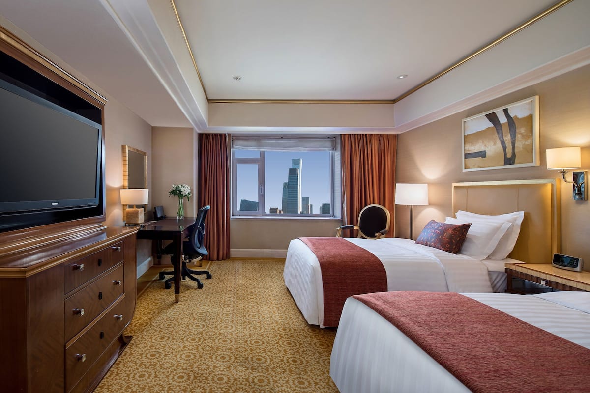 A hotel room with two beds, a desk, a TV, and a view of a cityscape.