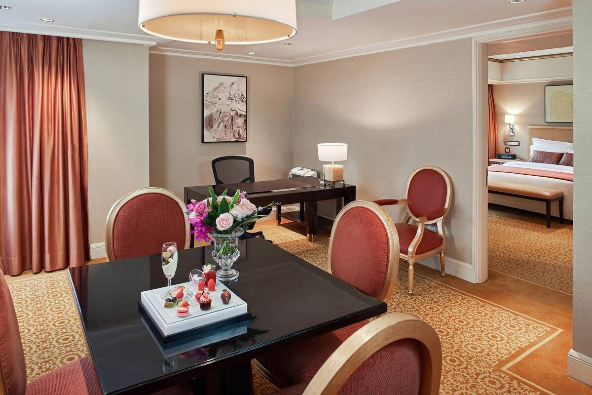 A hotel room with a dining table set for two, a bed, a desk with a lamp, and a painting on the wall.