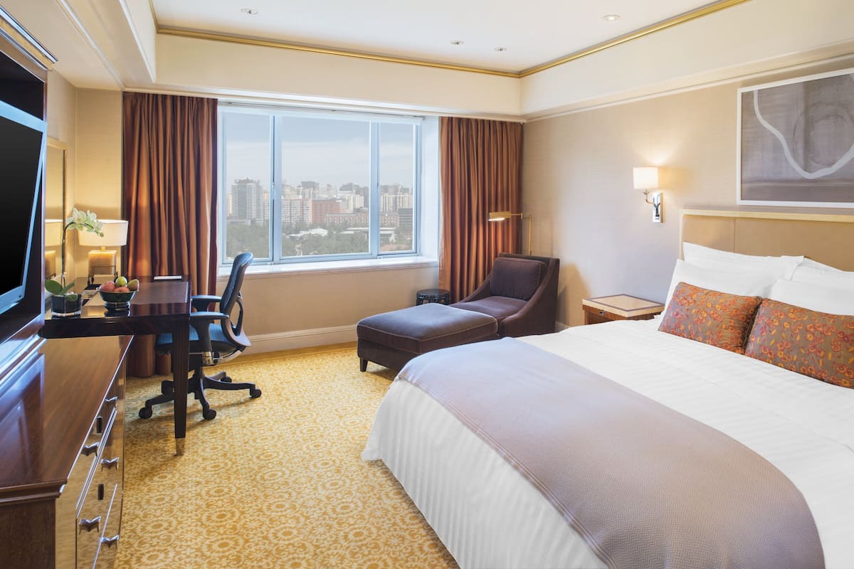 A hotel room with a large bed, a desk with a chair, a small table, a lamp, and a view of the city through the window.