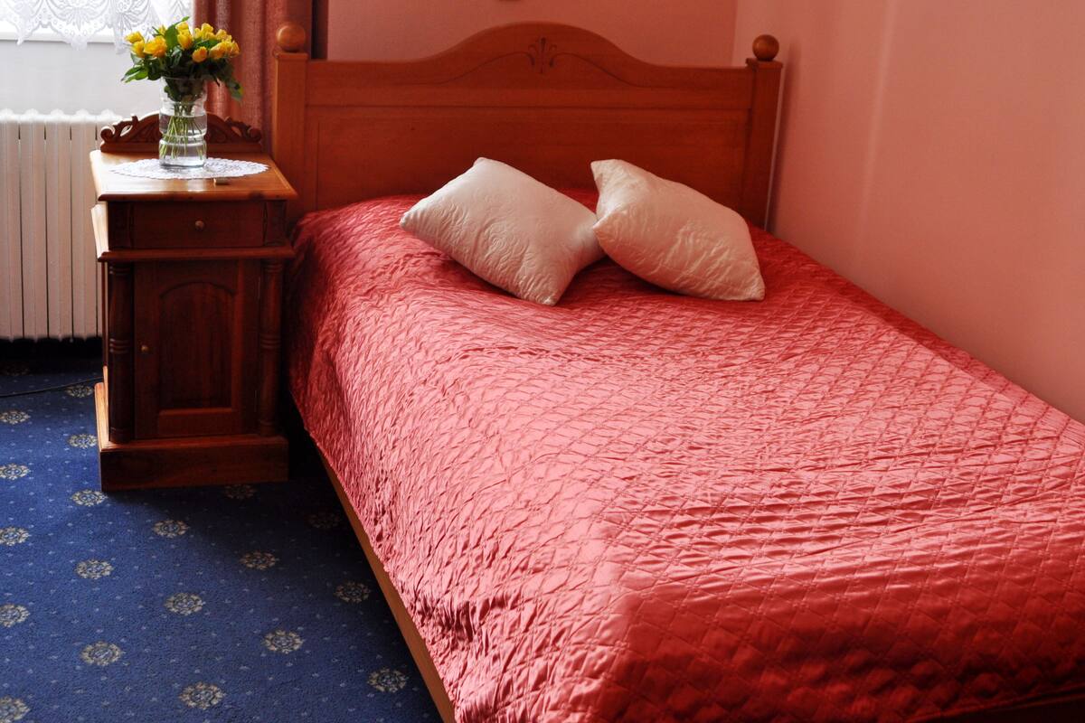 Quarto com cabeceira de madeira, colcha de cama acolchoada vermelha, duas almofadas brancas, mesa de cabeceira de madeira com um vaso de flores amarelas e tapete azul estampado.