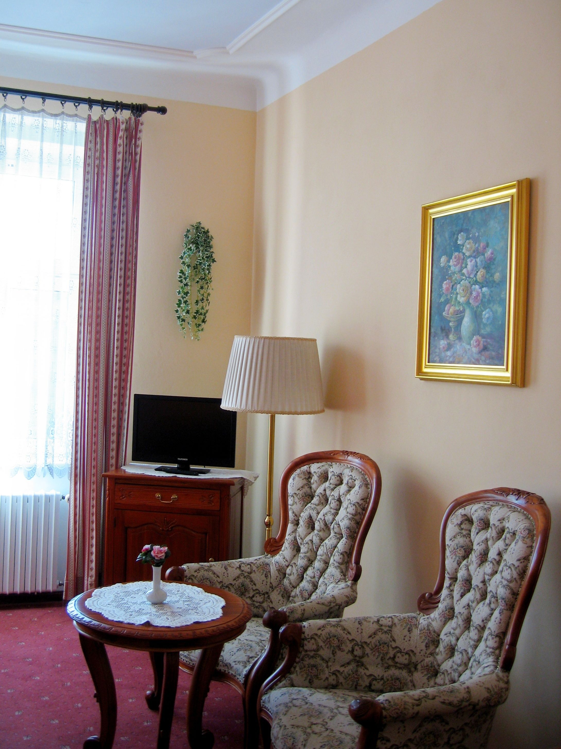 A room with two upholstered chairs, a small round table with a lace doily, a television on a wooden cabinet, a floor lamp, a framed painting, and a window with curtains.