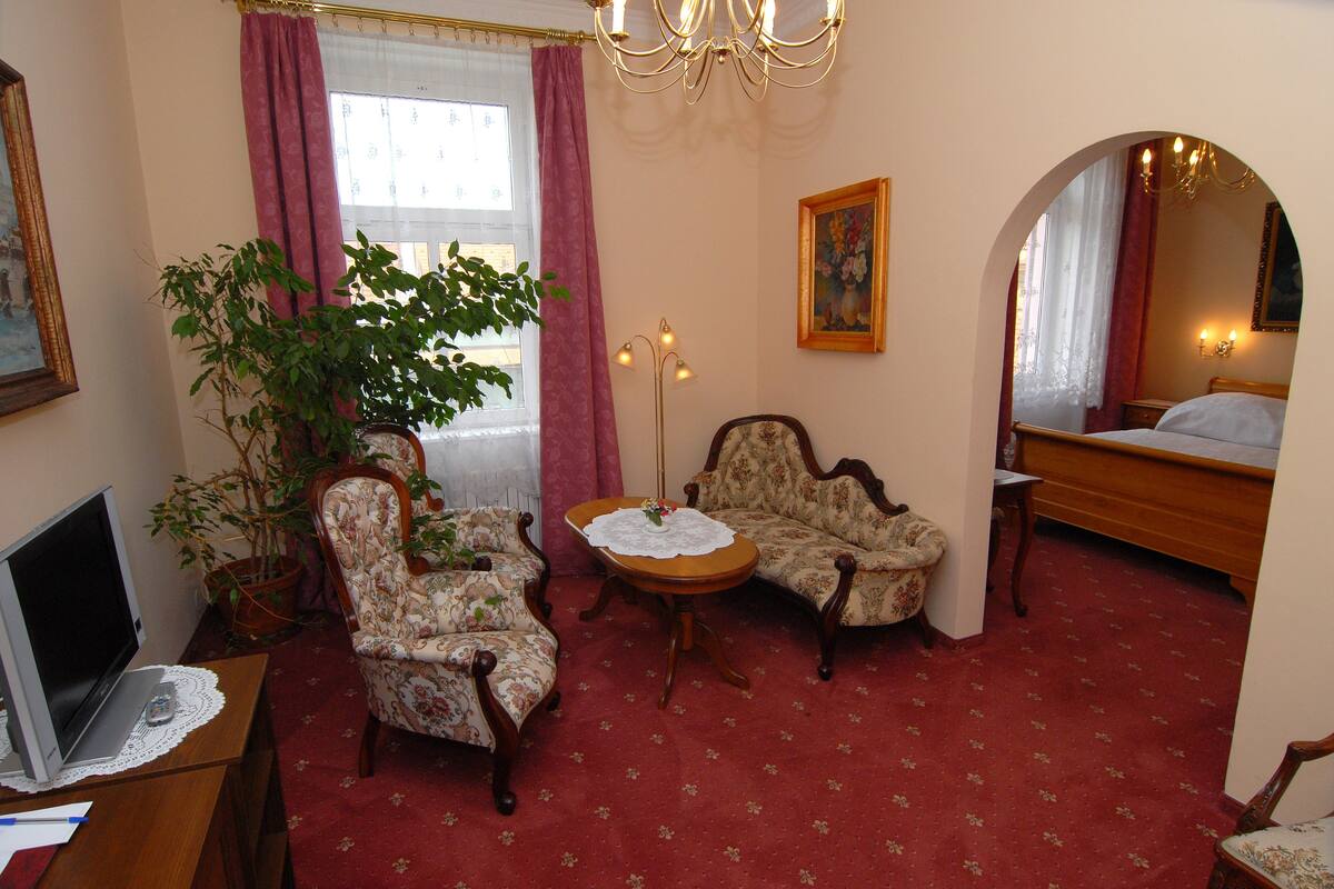 Quarto de hotel com uma cama, duas poltronas, uma mesinha, um vaso de planta, uma escrivaninha com um computador e um espelho grande.